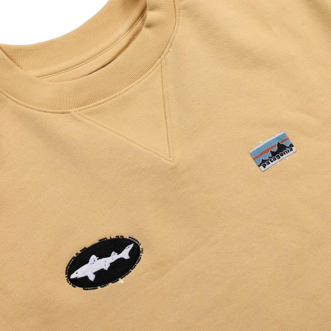 A close-up of the Patagonia Tan Crewneck Sweatshirt featuring two patches: an oval shark patch and a rectangular mountain logo with "Patagonia.