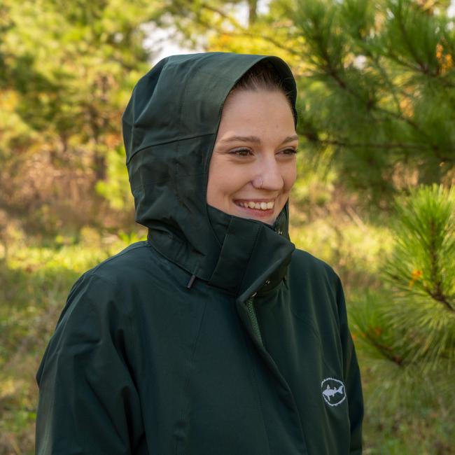 A smiling person wears the Patagonia W Dark Green 3-in-1 Parka by Patagonia while standing outdoors in a forested area surrounded by pine trees and greenery.