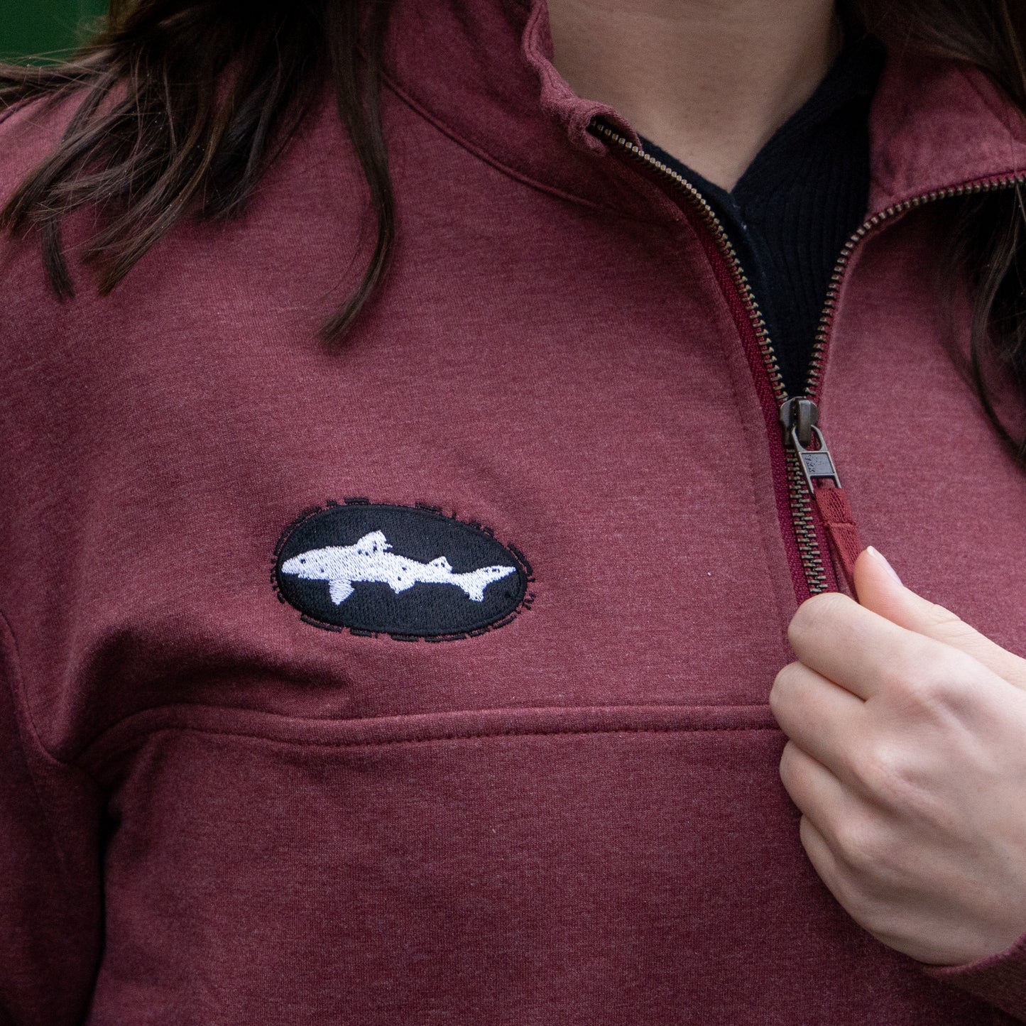 A person wearing the Patagonia W Deep Red Ahnya Quarter Zip Pullover, featuring a black and white fish-shaped chest patch, is slightly unzipping it with their right hand.