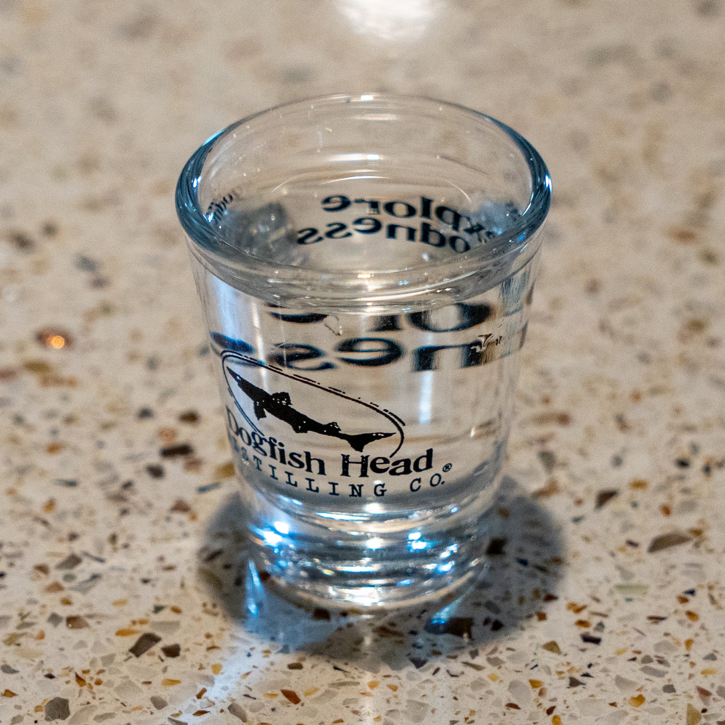 A 2.5 oz clear Shot Glass from Grandstand Glassware & Apparel rests on a speckled countertop, filled with transparent liquid—ideal for savoring your favorite spirits in style.