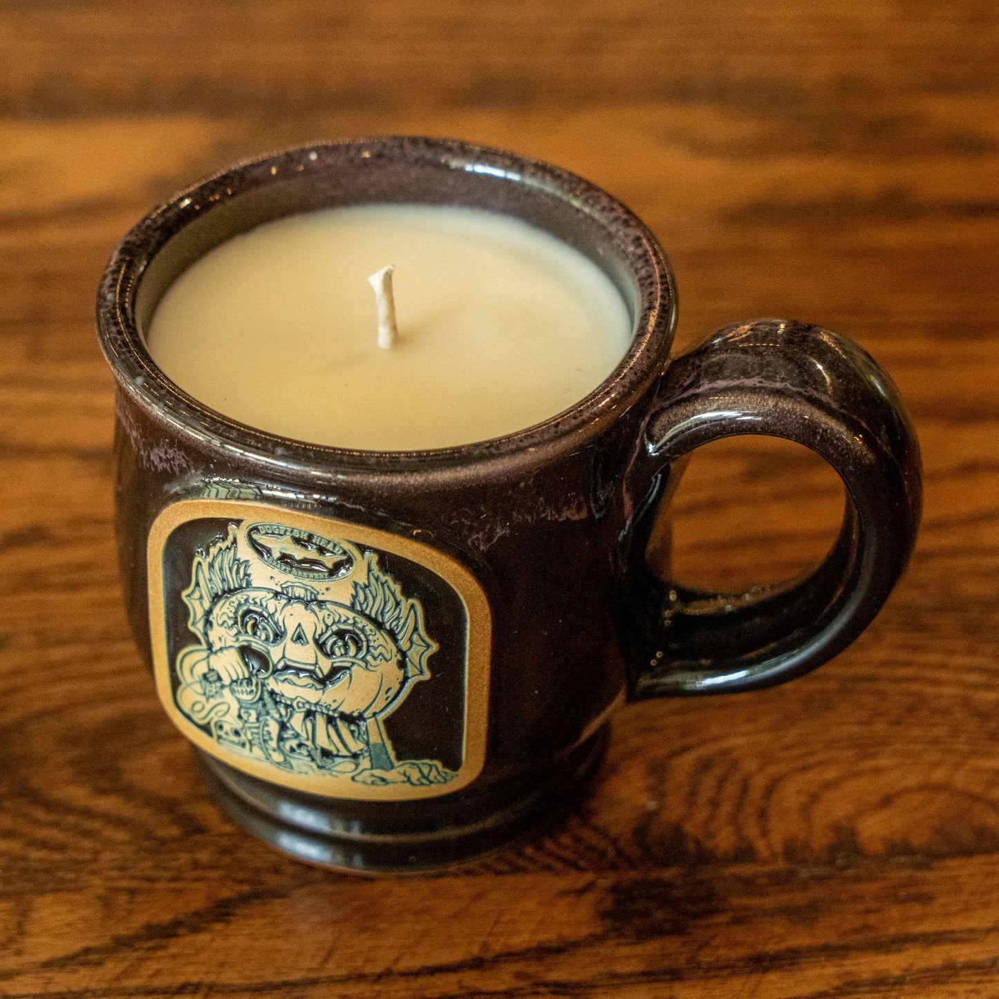 The Unwined Candles, LLC Punkin Mug Candle features a brown ceramic mug with a skull design, filled with white wax in a fall apple crumble scent, displayed on a wooden surface.