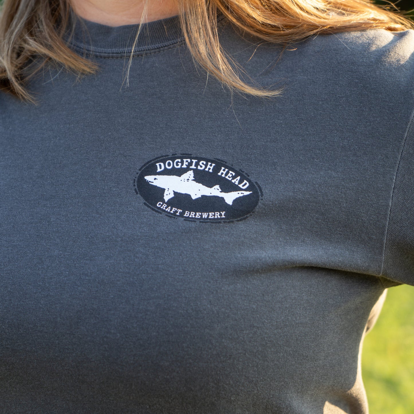 A person wearing the GO WEST T-SHIRT COMPANY Punkin Short Sleeve Tee, a garment-dyed dark gray shirt with a Dogfish Head Craft Brewery shark logo on the chest, stands outdoors with sunlight on their shoulder.