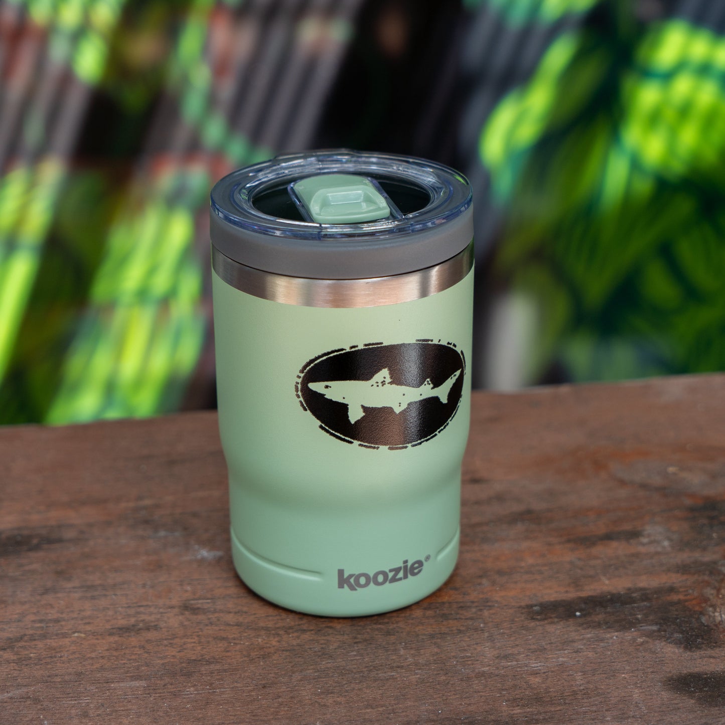 The Sage 12oz Koozie Can Insulator by Arrasmith Promotions LLC is a light green, vacuum-insulated stainless steel koozie with a lid and black and white shark logo, "koozie" printed near the base, shown on wood with a leafy blurred background.