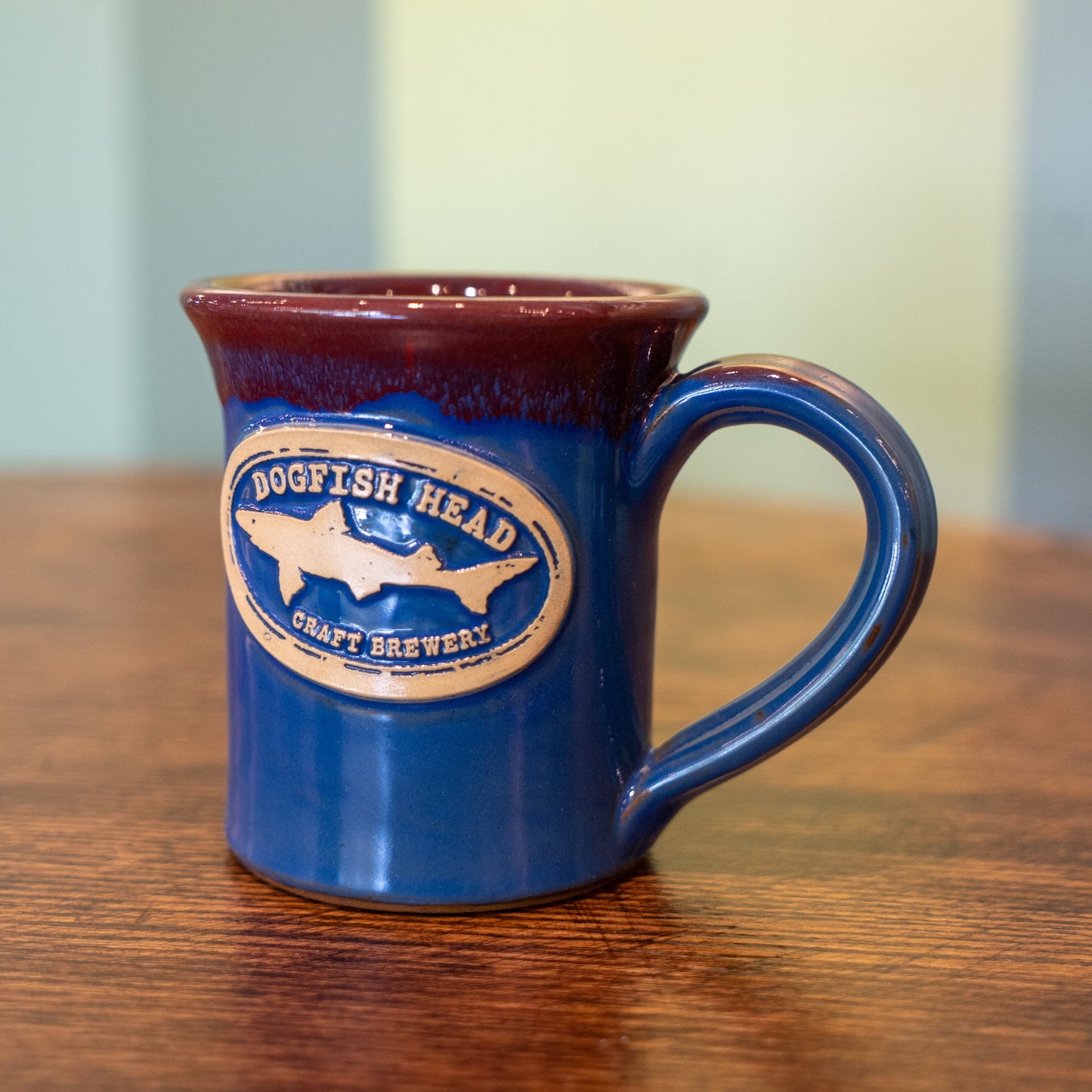 The Sky Blue Core Flare Ceramic Mug by Deneen Pottery features a maroon rim and beige oval logo with a dogfish image and "Dogfish Head Craft Brewery" text, shown sitting on a wooden table.