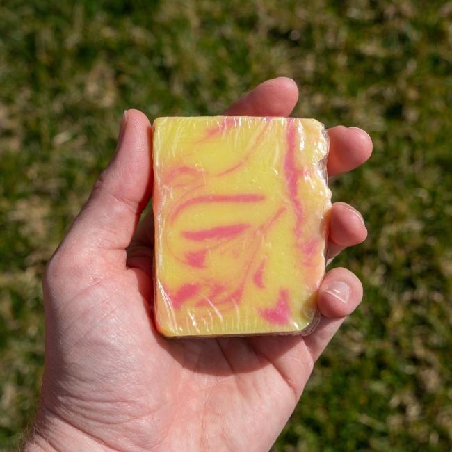 A hand holds an Indigo Bee Strawberry Lemonade Bar Soap with yellow and pink swirls, wrapped in clear plastic, against a blurred grassy background.