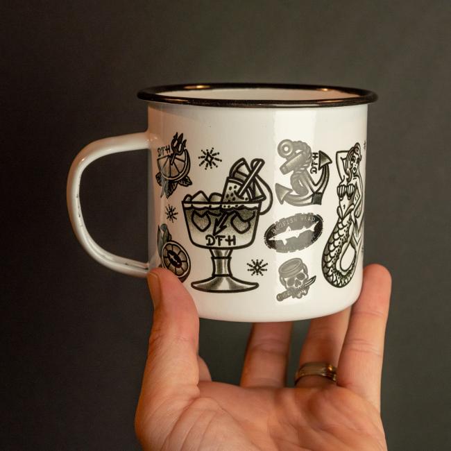 A hand holds the PROforma Tattoo Enamel Mug, featuring a black rim and quirky black tattoo-style designs including a mermaid, cocktail glass, cherries, and a snake.