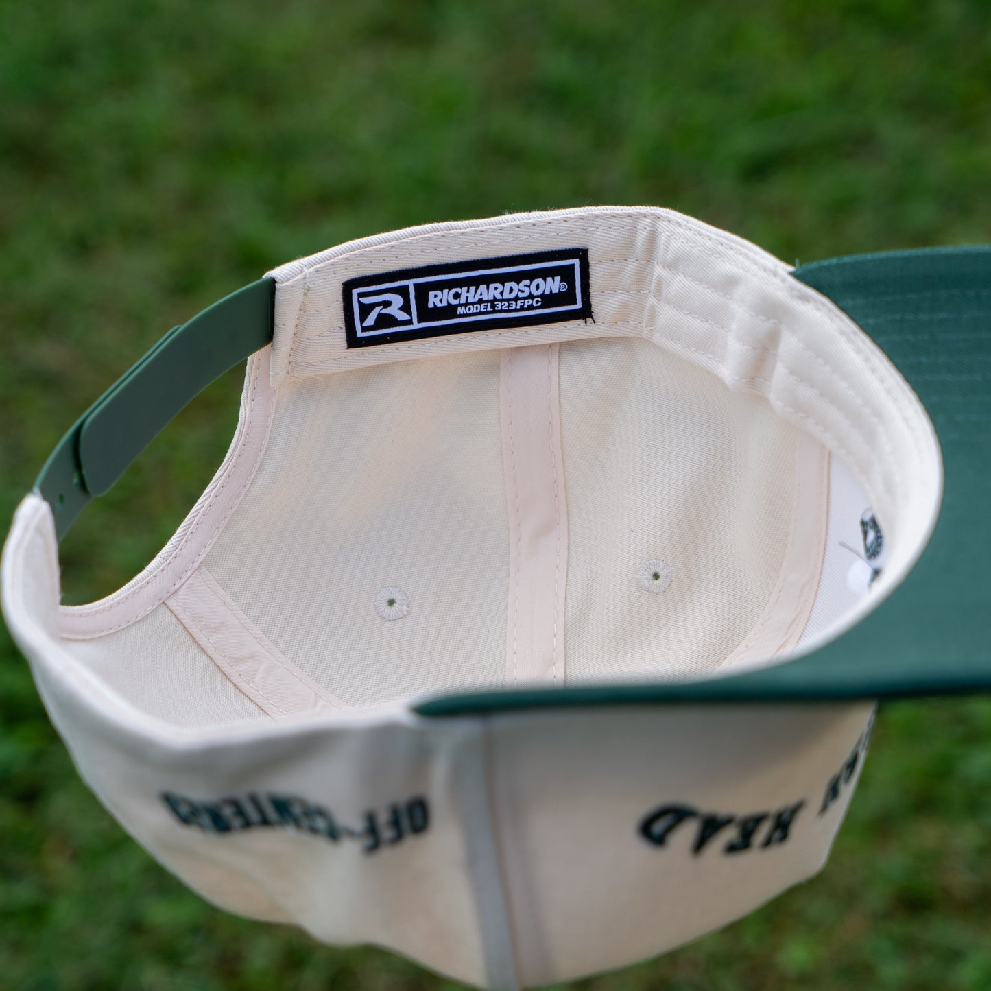 An open Upright DOGFISH Five Panel Snapback by Richardson in bold green and white is held upside down, revealing the inside label, with grass in the background—ideal for Dogfish Head fans.
