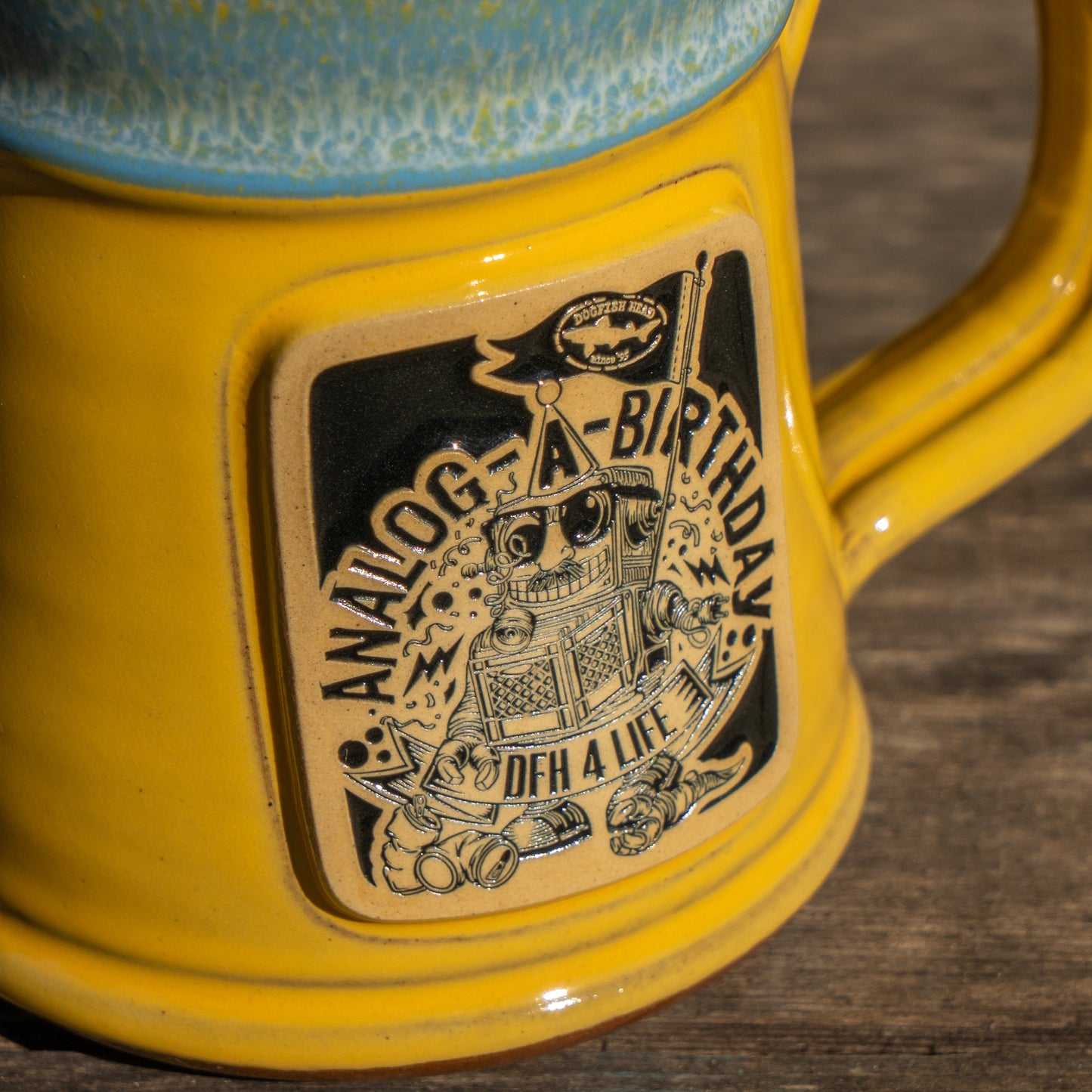 A close-up of the Yellow Analog Splitrock Ceramic Mug by Deneen Pottery, featuring a badge with a robot, flag, gears, and the text "ANALOG-A-BIRTHDAY" and "DFH 4 LIFE," shown on a wooden surface.