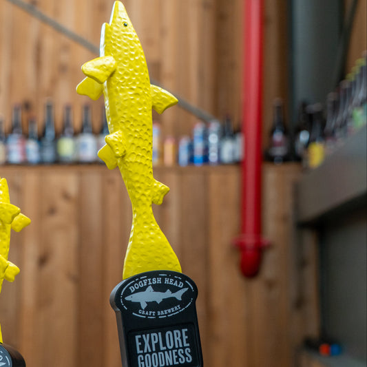 Two neon yellow shark-shaped Merchtank Yellow Shark Tap Handles stand before a wooden wall lined with bottles. One tap displays EXPLORE GOODNESS in bold white text.