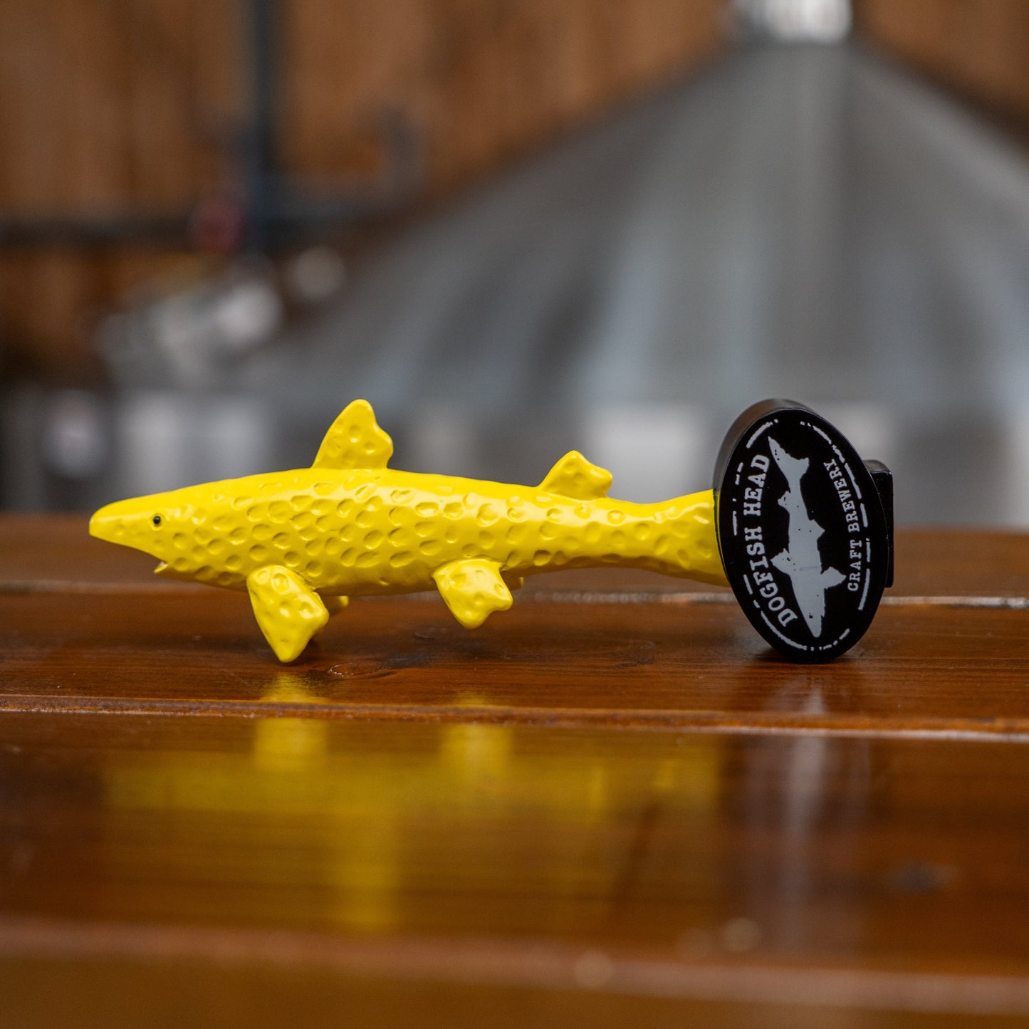 A neon yellow, textured shark figurine labeled Tap Handle - Shorty by Merchtank sits on a wooden surface, evoking the spirit of craft beer, with a blurred industrial background.