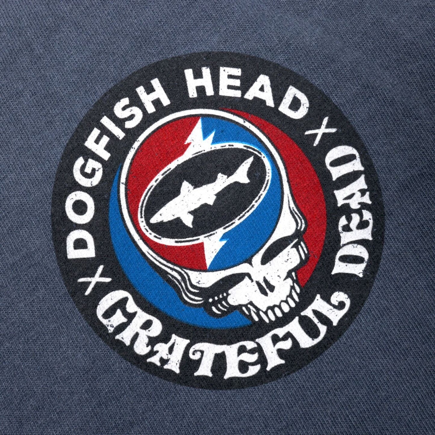Close-up of the collaboration logo which is a circle bordered in black with writing "Dogfish Head x Grateful Dead". The inside of the circle is half red and half blue with a white skull in the center. In the skulls head there is a white lightning bolt with the Dogfish Head shark and shield logo on it.