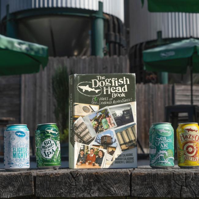 Four colorful Dogfish Head beer cans sit on a wooden surface beside the "26 Years of off Centered Adventures" book by J Wiley and Sons Publishing, with sunny outdoor brewery elements blurred in the background.