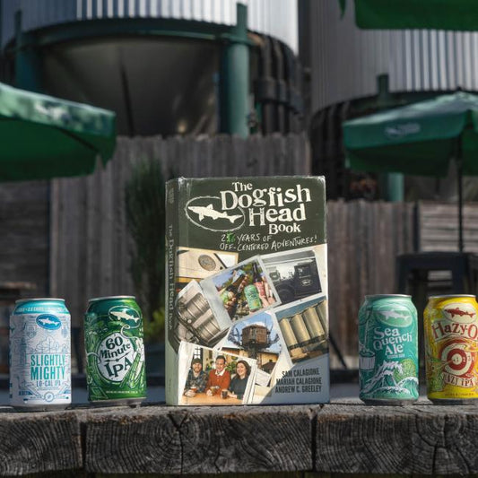 Four colorful Dogfish Head beer cans sit on a wooden surface beside the "26 Years of off Centered Adventures" book by J Wiley and Sons Publishing, with sunny outdoor brewery elements blurred in the background.