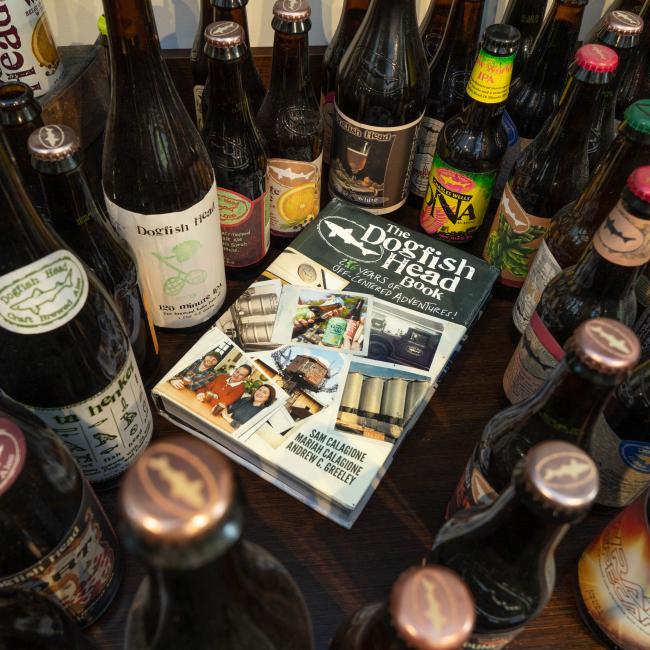 A selection of craft beer bottles encircles a copy of "26 Years of off Centered Adventures" by J Wiley and Sons Publishing, displayed on a wooden table with the cover featuring images and brewing gear.