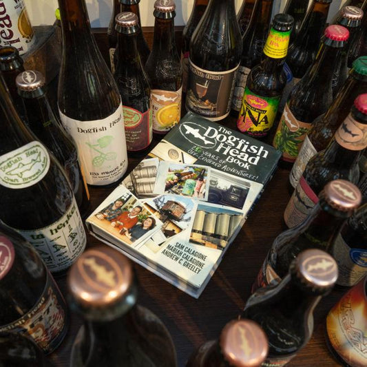Several Dogfish Head Craft Brewery beers are arranged around the "Dogfish Head Book 26 Years of off Centered Adventures" by J Wiley and Sons Publishing, capturing the innovative spirit of craft beer.