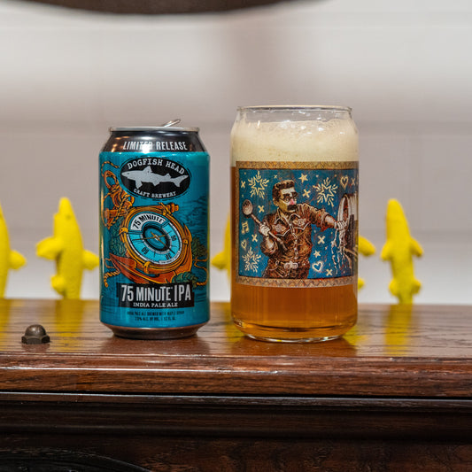 A Grandstand Glassware & Apparel 75 Minute Can Glass filled with frothy beer sits on a wooden surface, with four yellow banana-shaped figurines in the background.