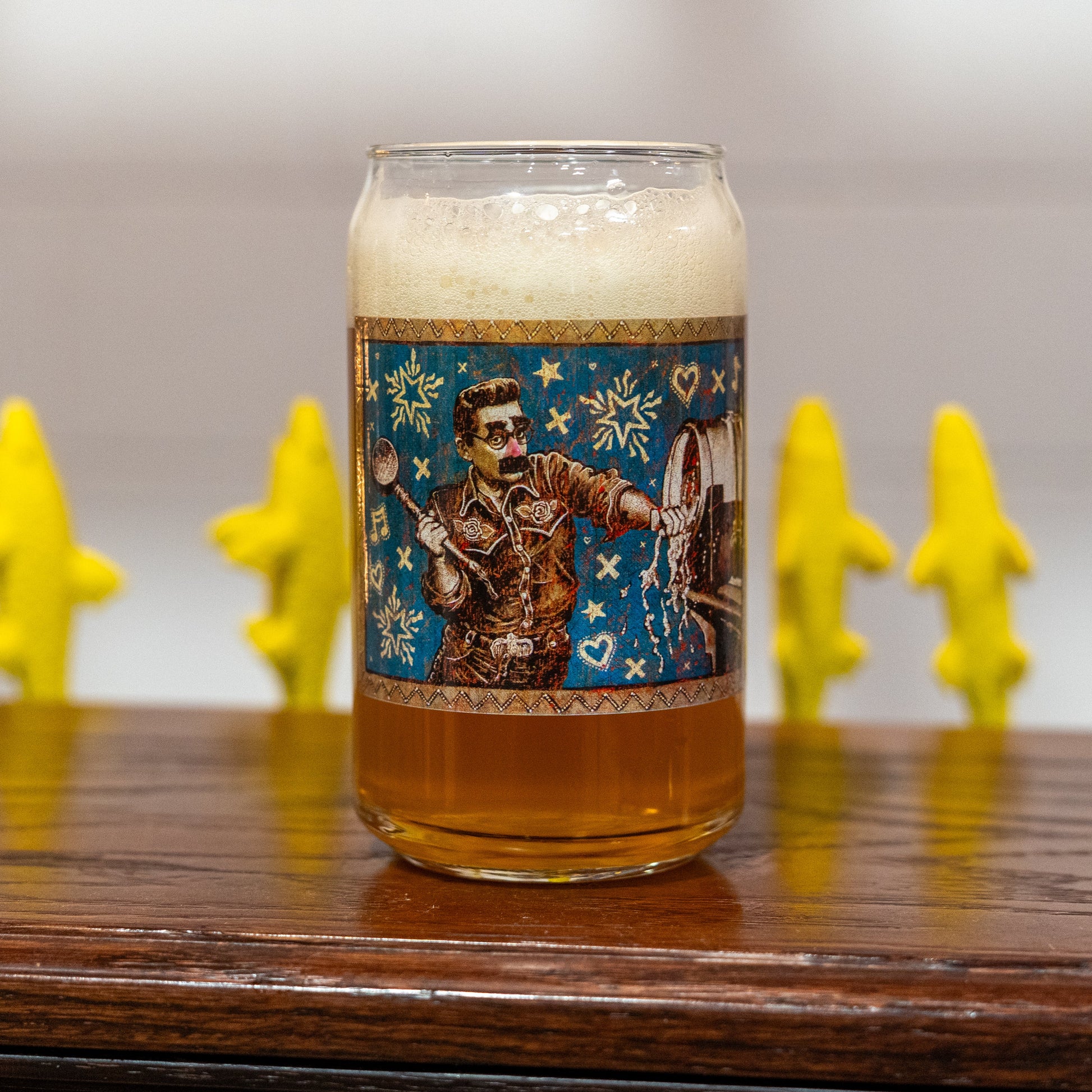 A 75 Minute Can Glass from Grandstand Glassware & Apparel, featuring colorful Elvis Presley artwork, sits on a wooden surface topped with foamy beer. Yellow alligator figures are visible in the blurred background.