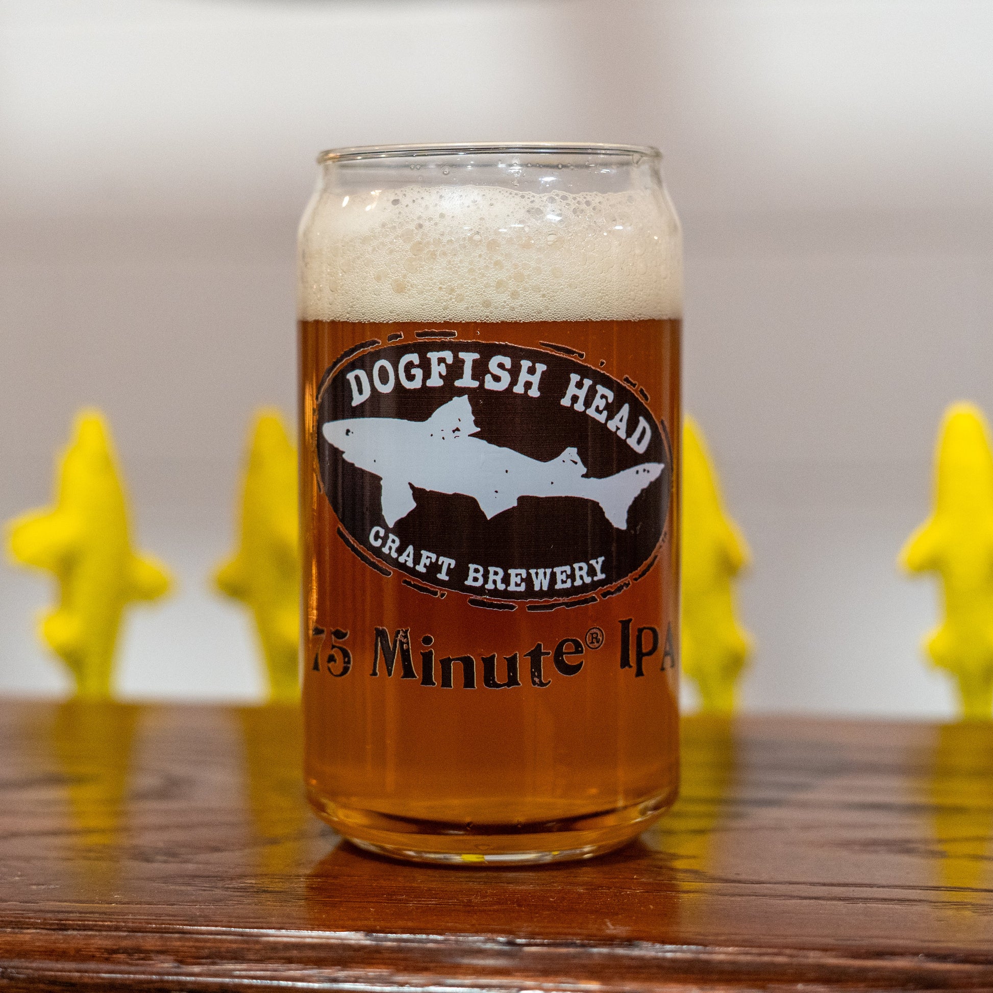 A glass of craft beer with a frothy head sits on a wooden surface, poured into the 75 Minute Can Glass by Grandstand Glassware & Apparel. The can glass displays the logo, with blurred yellow shark figurines in the background.
