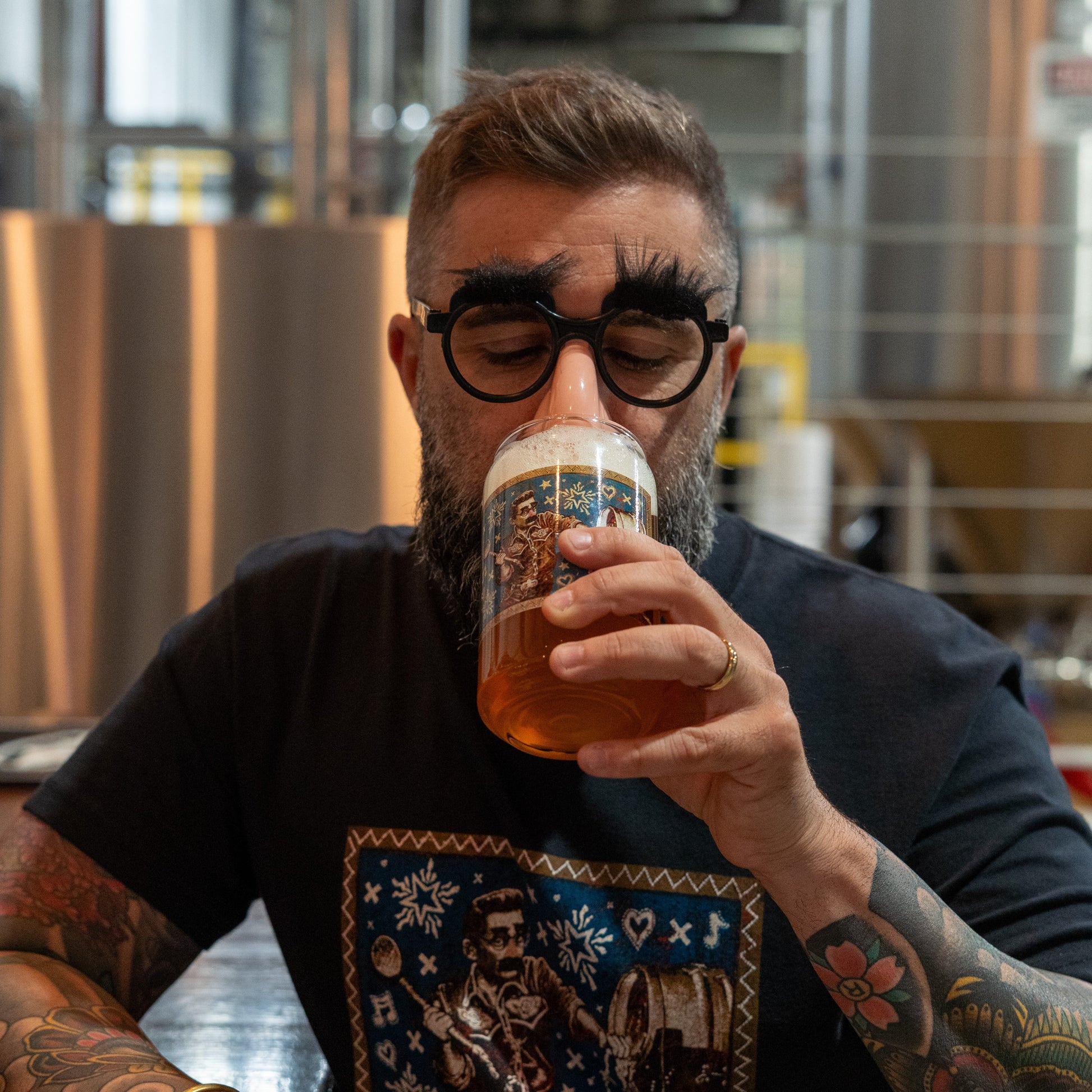 A tattooed man wearing a festive black t-shirt enjoys a pint from a Grandstand Glassware & Apparel 75 Minute Can Glass at the bar, with brewing equipment in the background setting the mood for this exclusive event.