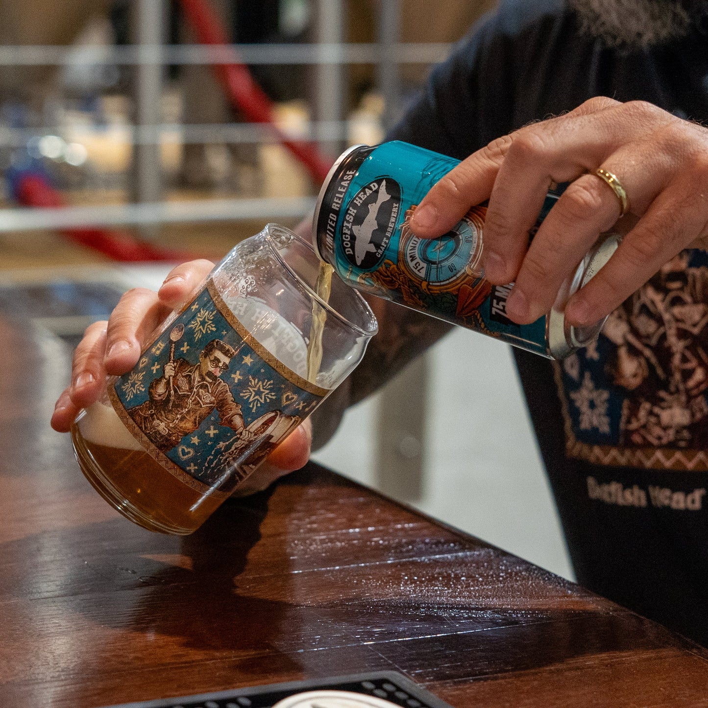 Someone pours Dogfish Head beer from a limited edition can into a 75 Minute Can Glass by Grandstand Glassware & Apparel on a wooden bar, wearing a black graphic shirt and gold ring.