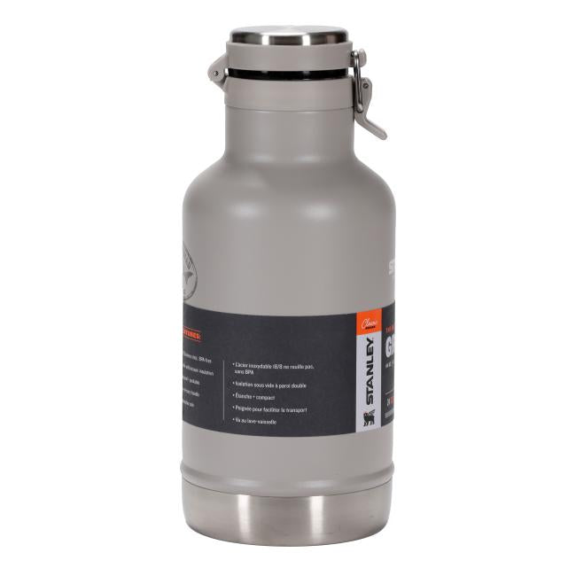 A light gray insulated growler with a stainless-steel base and top. It has a sturdy side handle that matches the growler's body. On the front, there are two labels: a smaller one on the left (partially obscured) and a larger, more prominent one on the right. The right label is black with an orange stripe down its left side, featuring the "STANLEY" logo vertically in white text, and an illustration of a bear.