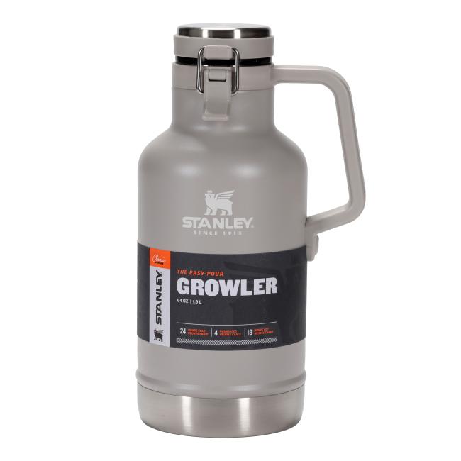 A light gray insulated growler with a stainless steel base and a matching stainless steel top with a gray handle. The handle is integrated into the cap and extends down the side of the growler. The front of the growler features a black label with an orange stripe on the left side. Above the black label, there is an embossed Stanley logo of a bear and a bottle, with "STANLEY." and "SINCE 1913" below it.