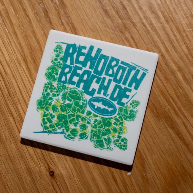 The B&E Coaster by Paint the Town, Inc. features "Rehoboth Beach, DE" in bold teal on an absorbent stone with green hops and a white shark logo, set against a wooden surface.