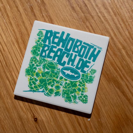 The B&E Coaster by Paint the Town, Inc. features "Rehoboth Beach, DE" in bold teal on an absorbent stone with green hops and a white shark logo, set against a wooden surface.