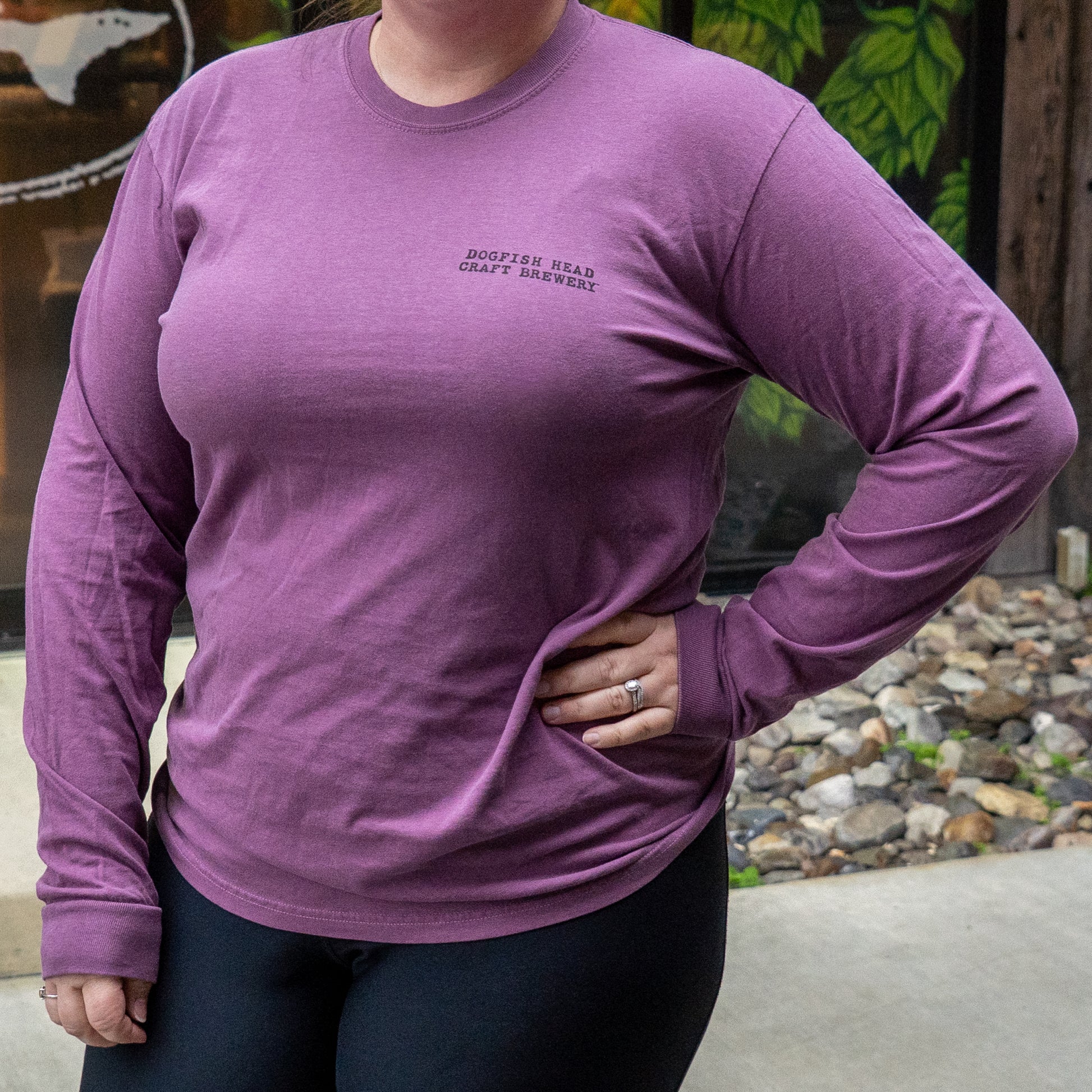 A person stands outdoors with one hand on their hip, wearing the GO WEST T-SHIRT COMPANY Berry Oversize Logo Long Sleeve shirt in mauve. The background includes plants, rocks, and a partial mural.