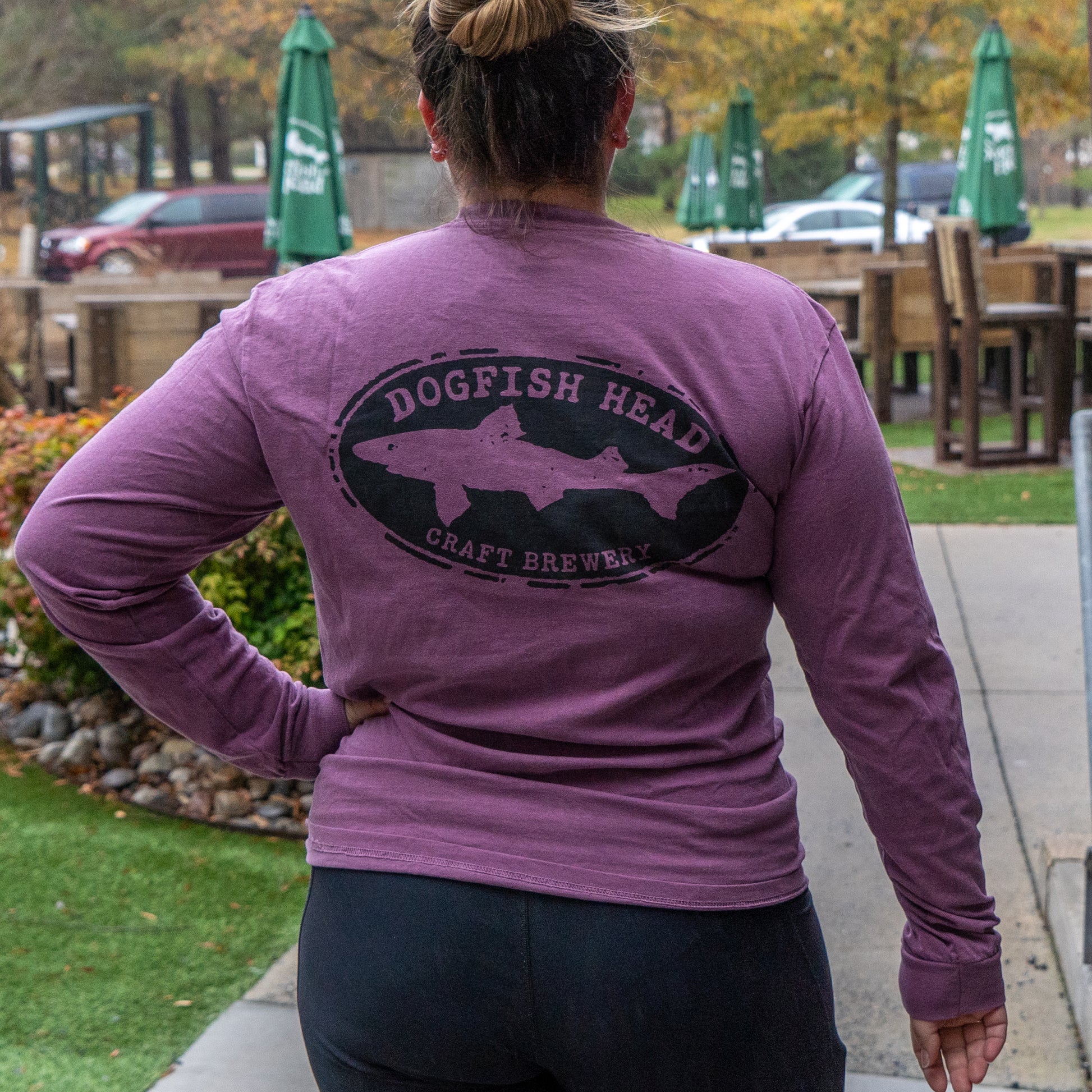 A person with their back to the camera wears a Berry Oversize Logo Long Sleeve by GO WEST T-SHIRT COMPANY and black leggings, standing outdoors near tables and green umbrellas on a cloudy day.
