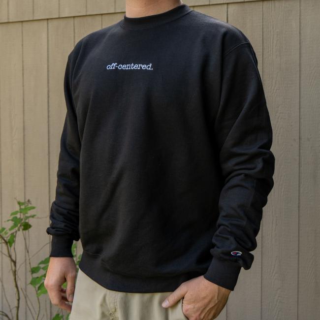A person in the GO WEST T-SHIRT COMPANY Black Off Centered Crew Sweatshirt, with "off-centered." in white on the chest, stands before a wooden fence. Only their upper body and part of the arms are visible.