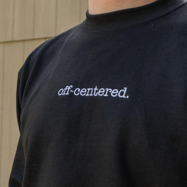 A person wears the GO WEST T-SHIRT COMPANY Black Off Centered Crew Sweatshirt, a cotton-polyester blend with "off-centered." embroidered in white on the chest, standing before beige wooden paneling.