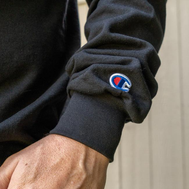 A close-up of a person's wrist and hand wearing a Black Off Centered Crew Sweatshirt by GO WEST T-SHIRT COMPANY, featuring the logo near the cuff. The background is blurred wood paneling.