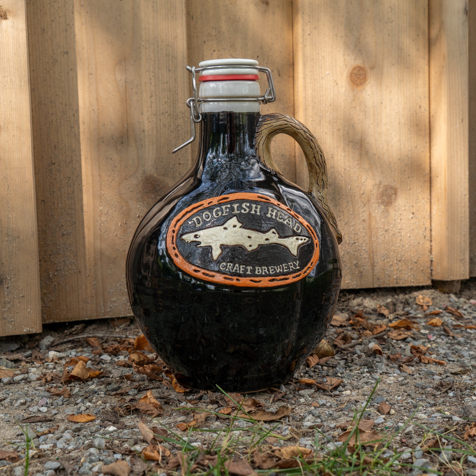 A Black Punkin Growler from Romanick Pottery, featuring a dark glass jug with a cork and latch closure, sits on the ground beside leaves and rocks in front of a wooden fence.