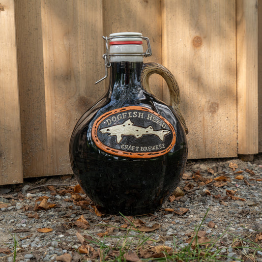 A Black Punkin Growler from Romanick Pottery, featuring a dark glass jug with a cork and latch closure, sits on the ground beside leaves and rocks in front of a wooden fence.