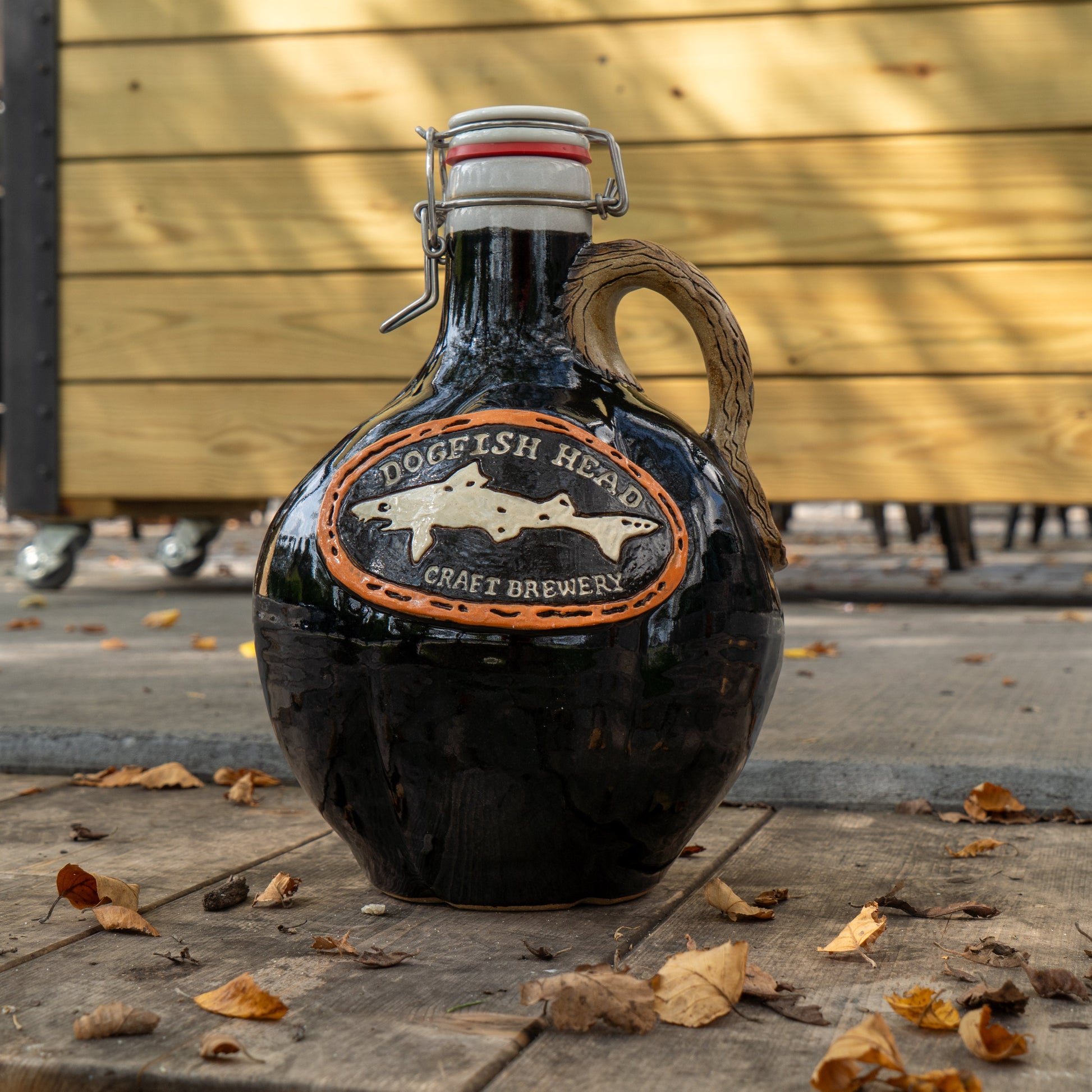 A Black Punkin Growler by Romanick Pottery with a handle and metal clasp sits outdoors on a wooden surface amid autumn leaves.