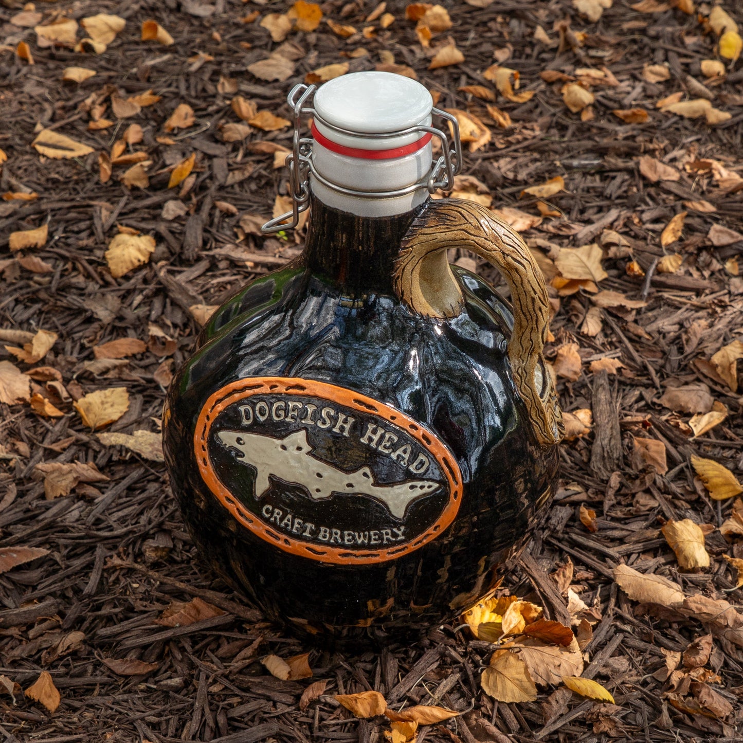 The Black Punkin Growler by Romanick Pottery, featuring a swing-top cap, rests on mulch among yellow and brown leaves.