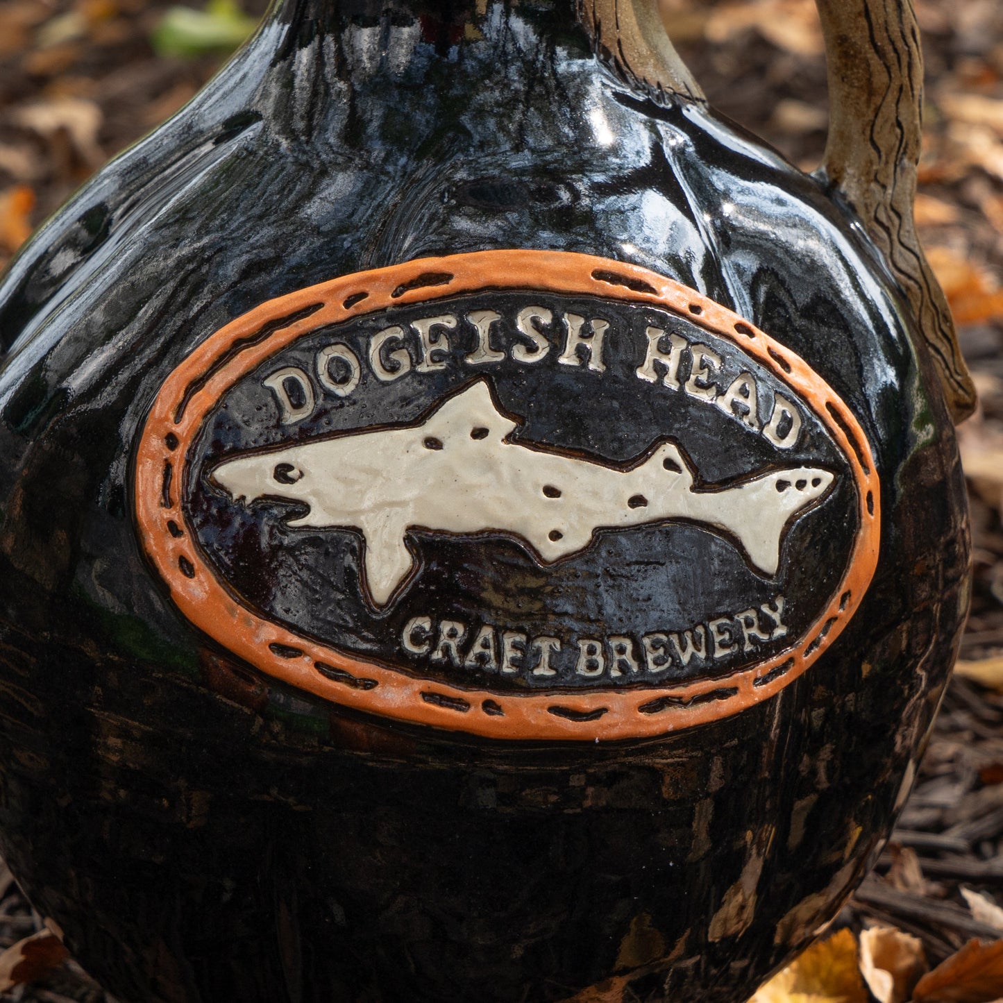 A close-up of the Black Punkin Growler by Romanick Pottery, a glossy black limited edition, showcases an oval plaque with a shark silhouette and raised lettering. Fallen leaves can be seen on the ground in the background.