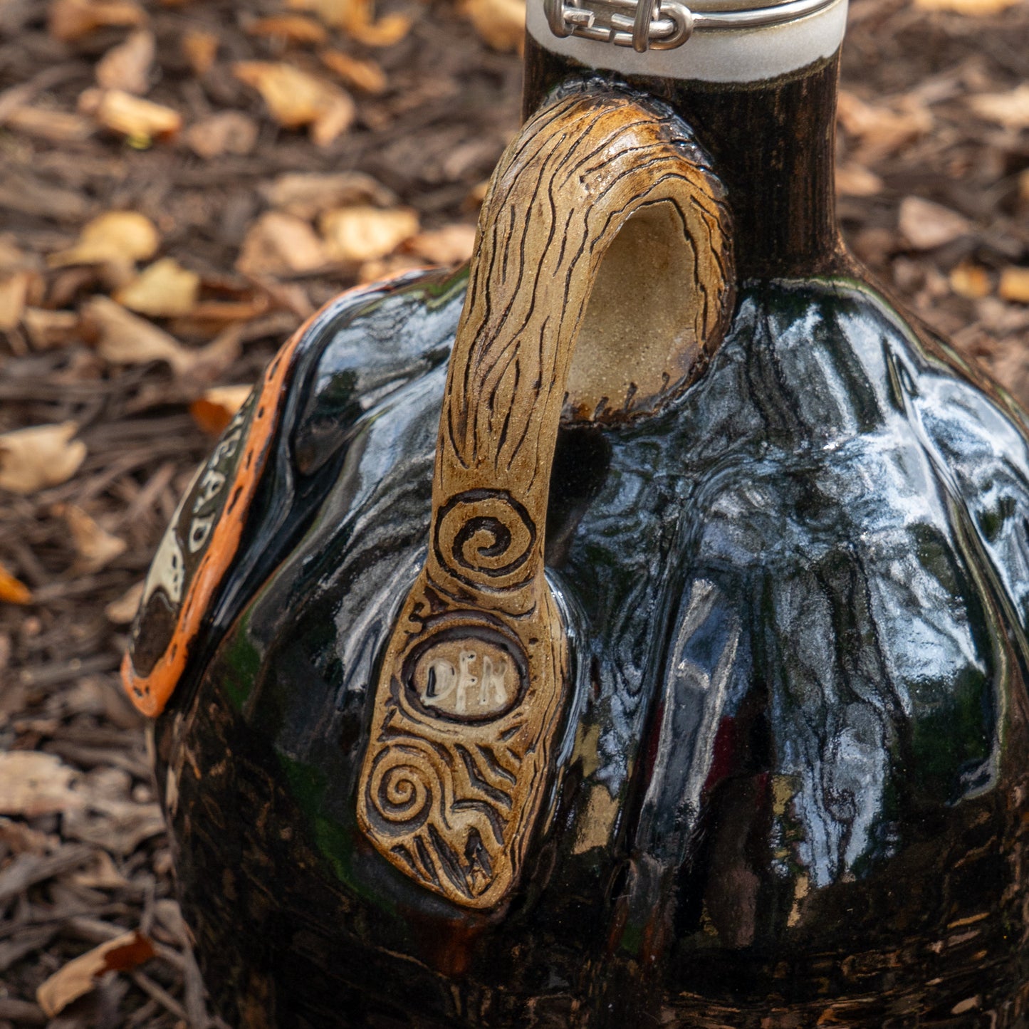 A close-up of the limited edition Black Punkin Growler by Romanick Pottery, featuring a dark glossy finish, textured carved handle, and engraved “DFN.” The handmade ceramic jug rests outdoors on brown mulch with scattered dry leaves.