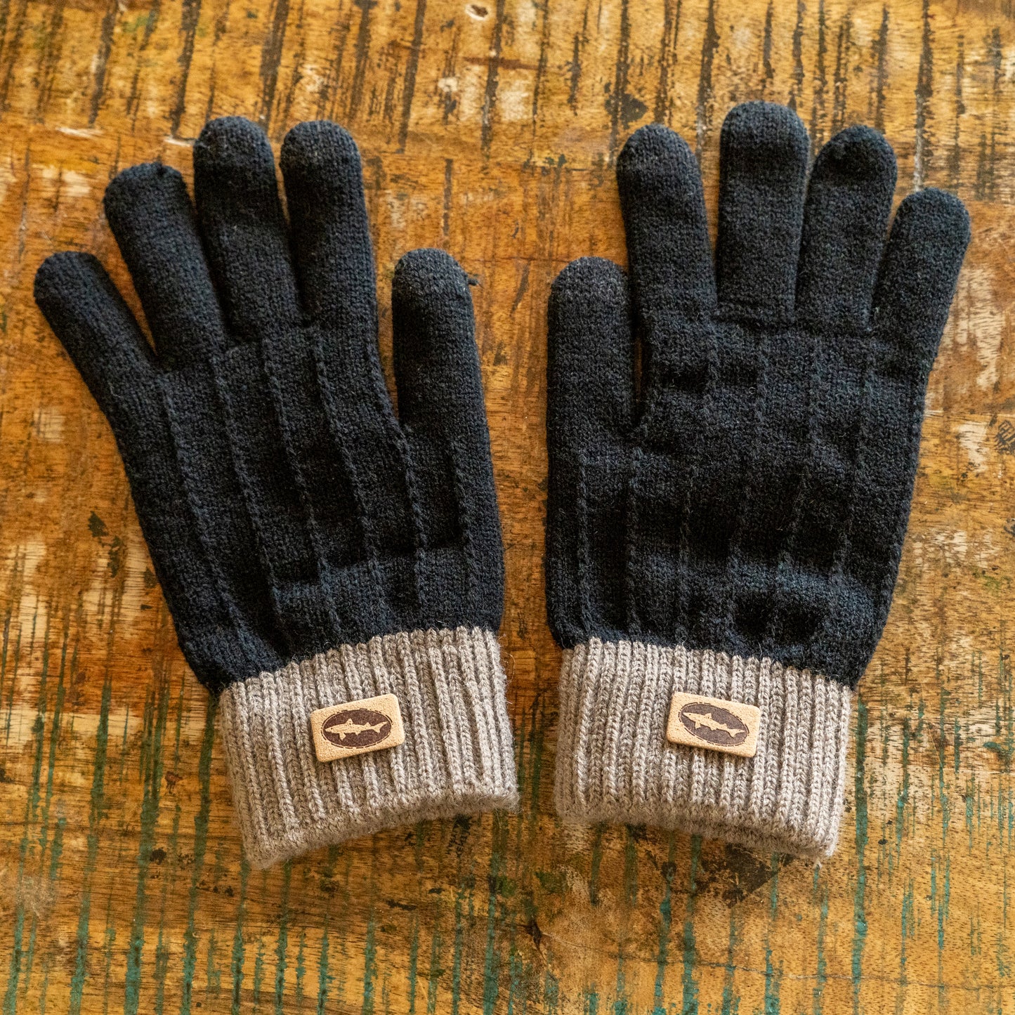 A pair of Black and Tan Gloves by Arrasmith Promotions LLC with gray ribbed cuffs and small brown fish logo patches, displayed on a rustic wooden surface.