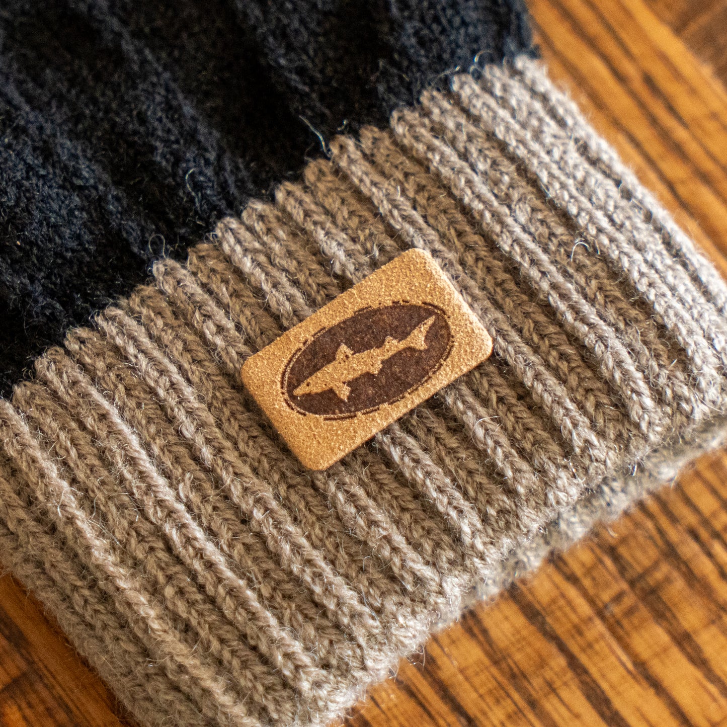 A close-up of a knitted beanie with black and gray stripes, featuring a rectangular leather patch with a fish logo, rests on wood—perfectly paired with Arrasmith Promotions LLC's Black and Tan Gloves.