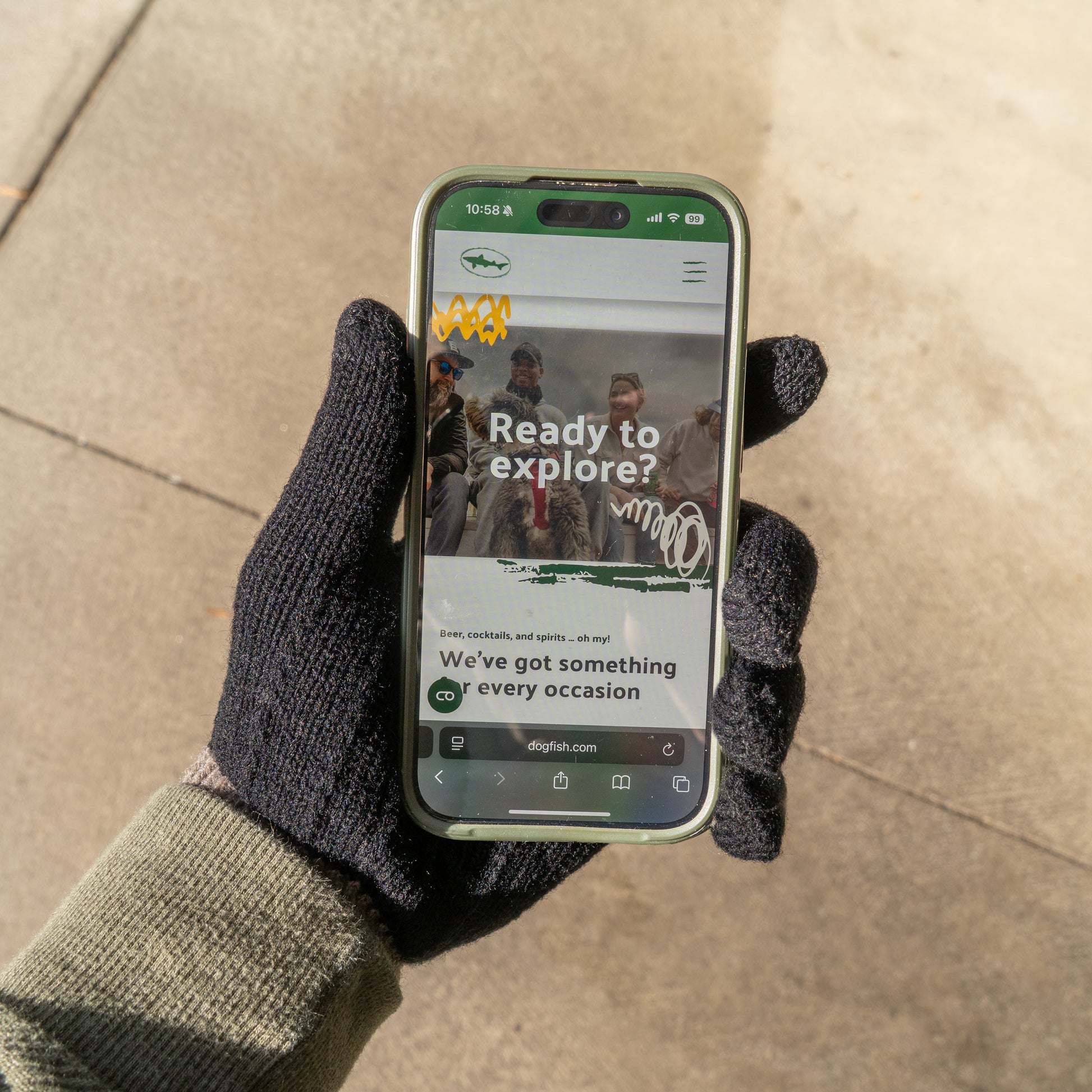 Someone wearing Arrasmith Promotions LLC’s Black and Tan Gloves, with a green sleeve visible, holds a smartphone showing an outdoor adventure site titled “Ready to explore?” with hikers pictured on the screen.