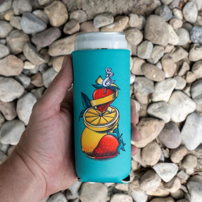 A hand holds a 12oz can in the Merchtank Blue Lemonade Slim Can Cooler, featuring turquoise color and artwork of a lemon, strawberries, and a mouse sitting on top, with rocks in the background.