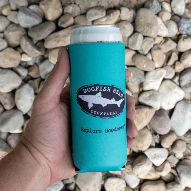A hand holds a Blue Lemonade Slim Can Cooler by Merchtank with a turquoise koozie, set against light-colored rocks.