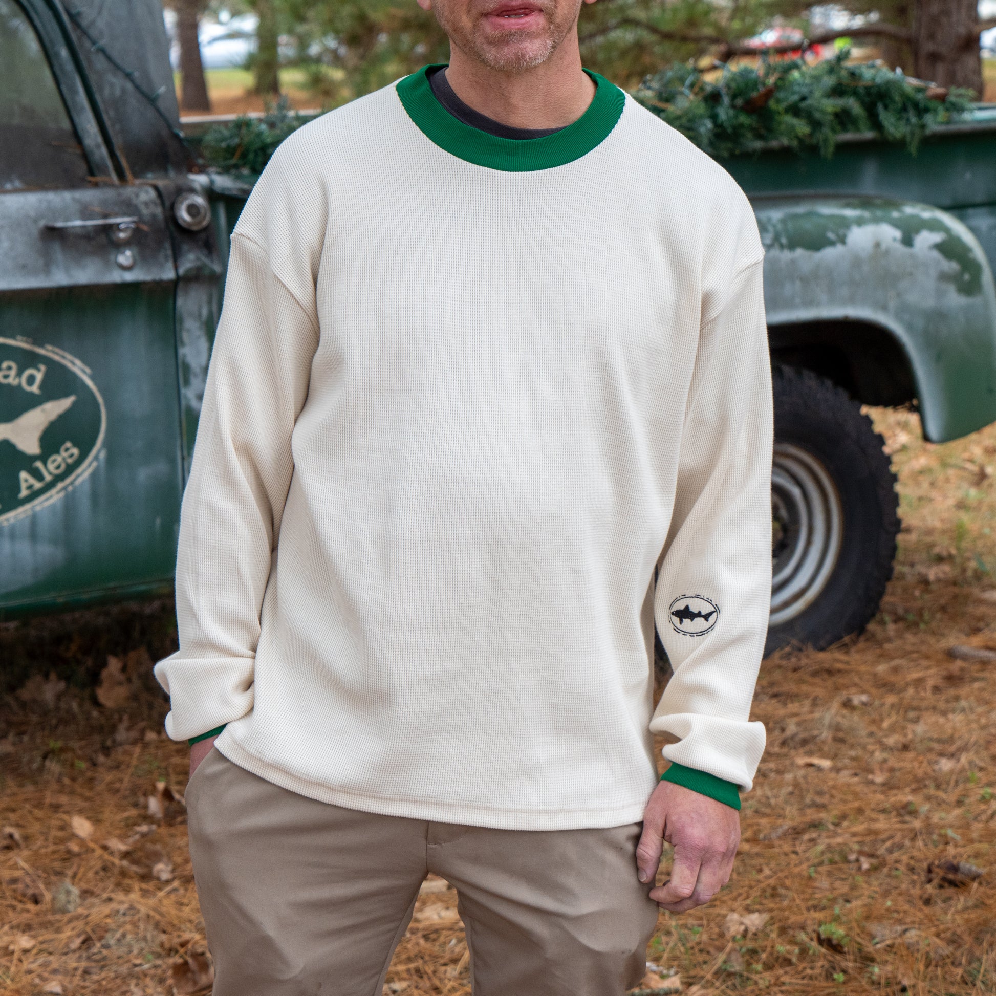 A man stands outdoors by an old pickup truck wearing the Dogfish Head x Boathouse Thermal Long Sleeve by Boathouse Apparel, a cream shirt with green collar and cuffs, a small sleeve logo, and pine needles scattered on the ground.