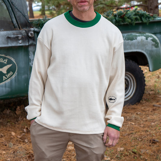 A man stands outdoors by an old pickup truck wearing the Dogfish Head x Boathouse Thermal Long Sleeve by Boathouse Apparel, a cream shirt with green collar and cuffs, a small sleeve logo, and pine needles scattered on the ground.