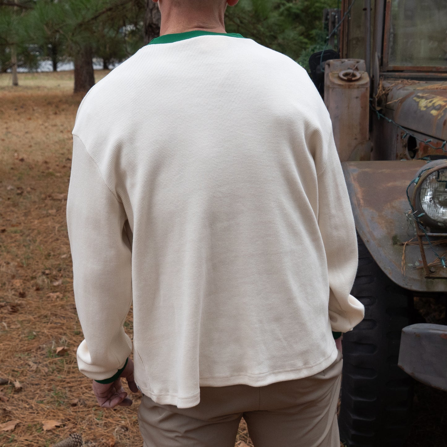 Someone wearing the Boathouse Apparel Dogfish Head x Boathouse Thermal Long Sleeve stands outdoors with their back to the camera beside a rusty old vehicle on pine needle-covered ground.