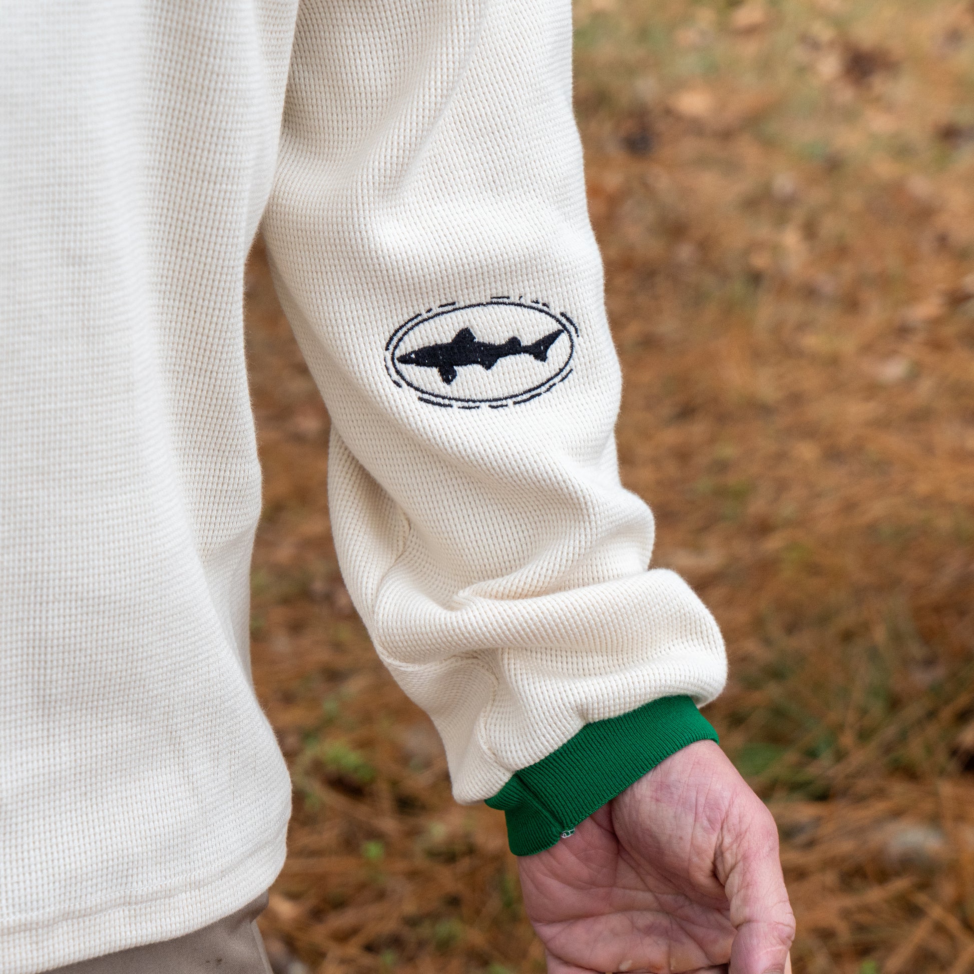 Someone wearing the Dogfish Head x Boathouse Thermal Long Sleeve by Boathouse Apparel, featuring a black shark logo on the sleeve and green cuffs, stands outdoors on brown grass.