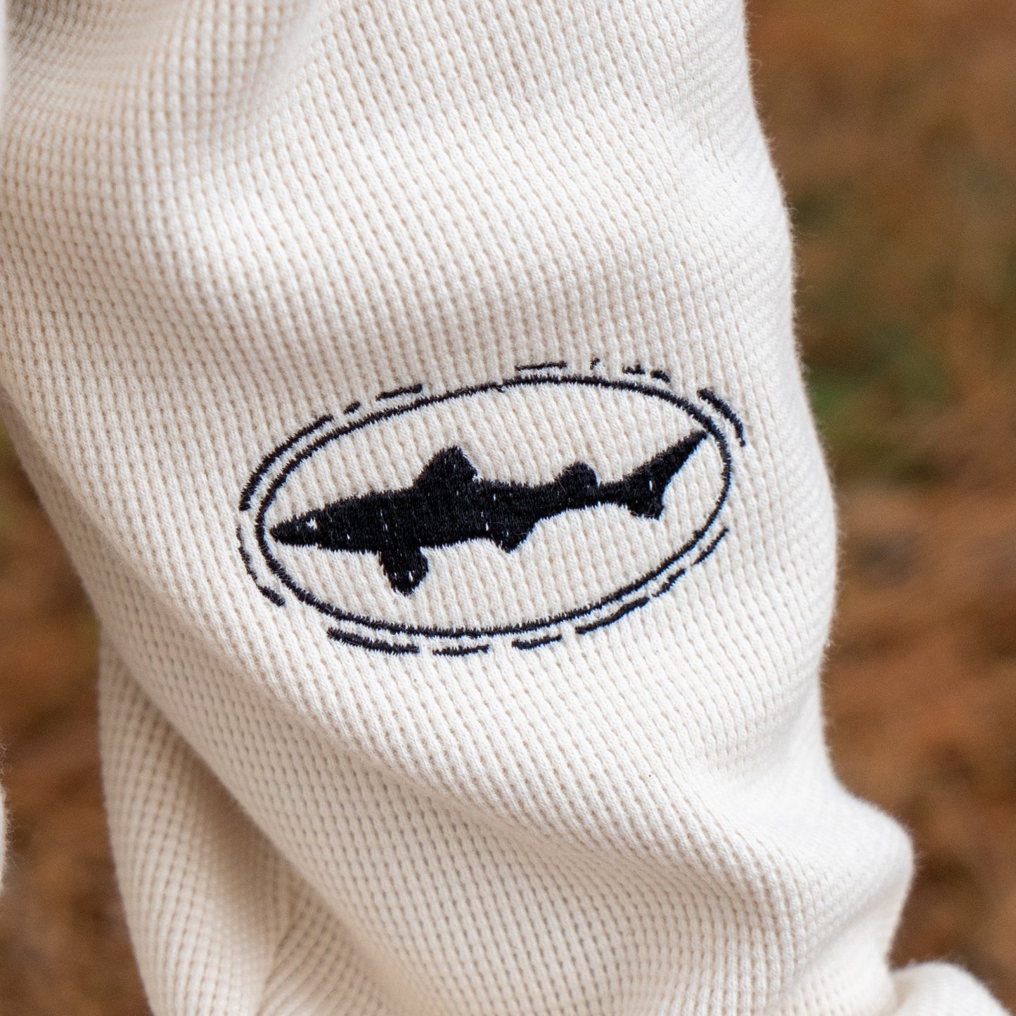 Close-up of cream-colored corduroy fabric on the Dogfish Head x Boathouse Thermal Long Sleeve by Boathouse Apparel, featuring a black embroidered shark outline inside an oval border. The background appears blurred.