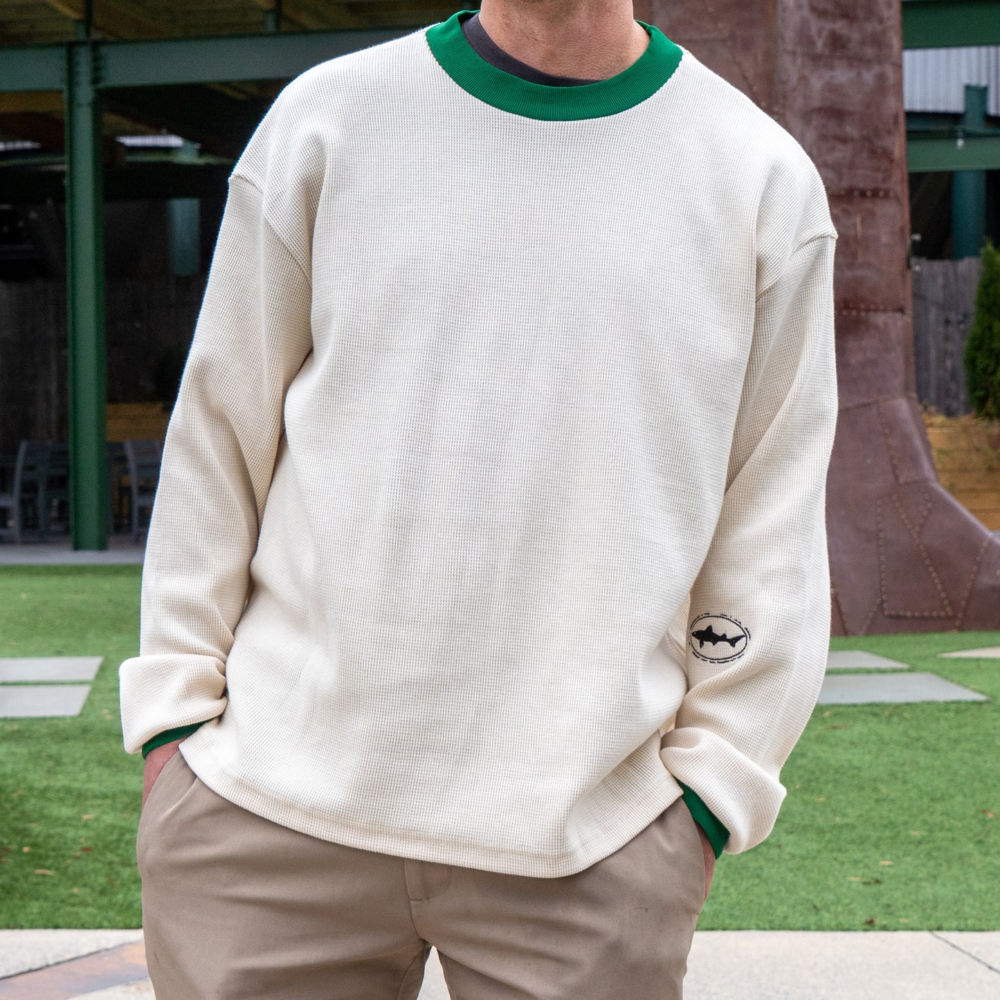 Wearing the Boathouse Apparel Dogfish Head x Boathouse Thermal Long Sleeve—cream with green collar—and khaki pants, a person stands outdoors on artificial grass with hands in pockets. A shark logo appears near the shirt’s hem.